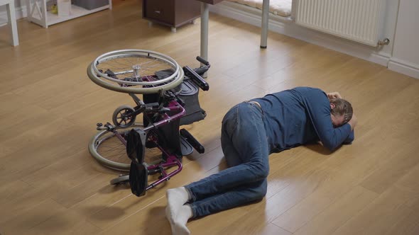 High Angle Wide Shot Desperate Crying Disabled Man Lying on Floor with Wheelchair Rolled Over alt