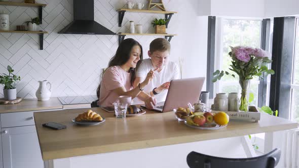 Couple Uses Laptop at Wooden Table in Brightly Lit Kitchen