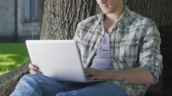 Young Man Sitting Under Tree Using Laptop, University Student Working on Project alt