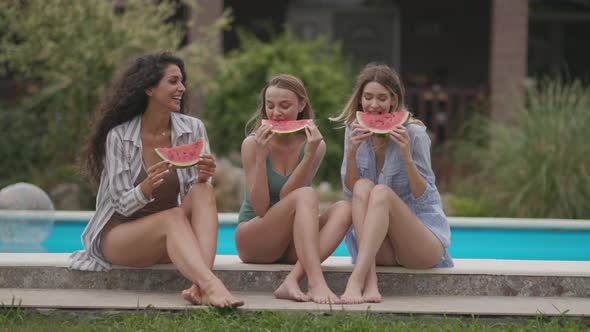 Three cute young women sitting on by the swimming pool and eating watermelon in the house backyard