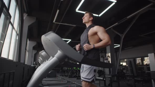 Athletic Man with Headphones is Jogging in Gym on Treadmill Side View alt