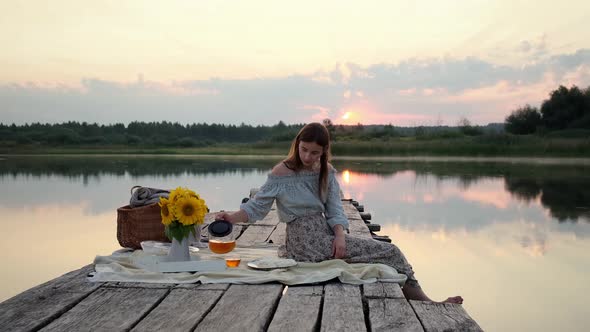 Happy Girl on the Pier at the Morning Sunrise on the Lake. Tasting Hot Tea and Sweets