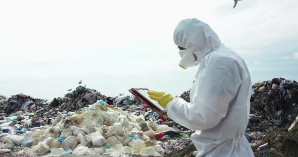 Man in Protective Suit and Gas Mask Noting in His Tablet at the Dump Landfill alt