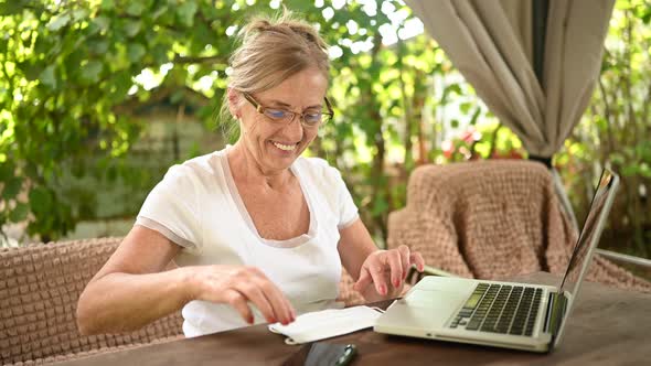 Elderly happy senior woman puts on protective face mask working online with laptop computer outdoor 