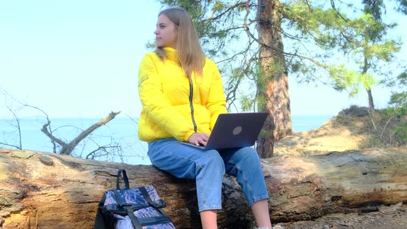 Attractive Student Uses a Laptop Sitting on a Log in the Woods alt