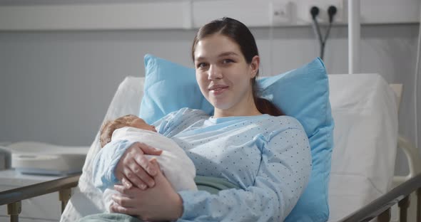Happy Young Mother Sitting in Hospital Bed Holding Newborn Baby Swaddled in Blanket alt
