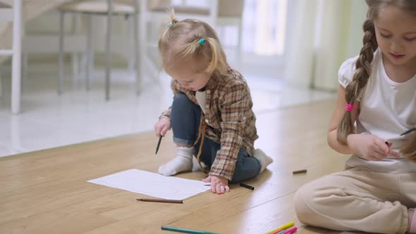 Cute Charming Little Blond Caucasian Girl Drawing Indoors with Blurred Brunette Sister alt