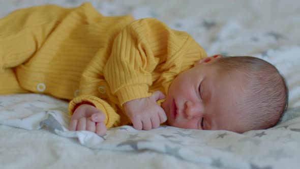 Small Cute Newborn Baby in Yellow Jumpsuit Lies on Side on Bed and Smiles alt