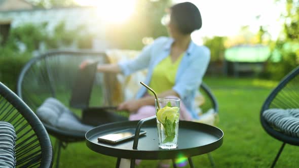 Woman Puts Down the Laptop and Takes Lemonade Sitting on Chair at Backyard