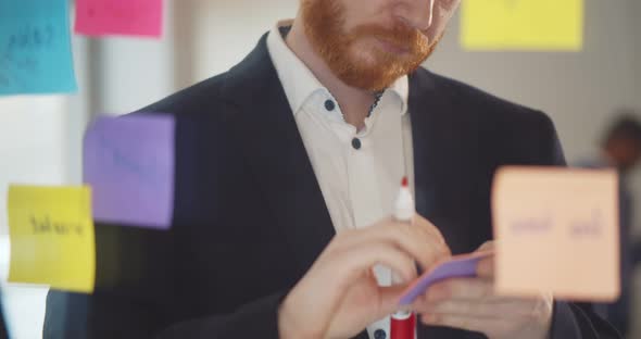Focused Businessman Writing on Sticky Notes and Attaching Them To Glass Wall alt