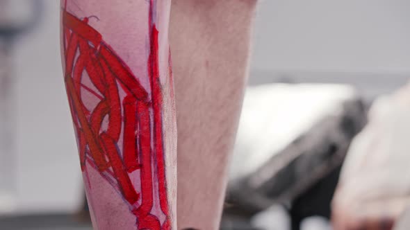 Tattoo Artist Adding Finishing Touches to a Sketch with Letters on the Leg on His Client with a Pen alt