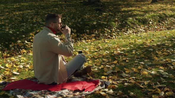 a Young Man is Sitting on a Plaid Blanket in an Autumn Park and Thoughtfully Drinks Tea From a alt