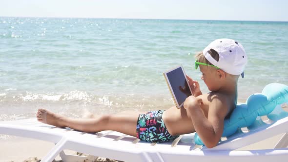 Cute Boy Is Lying on a Sun Lounger By the Sea and Playing Tablet Computer on a Sunny Clear Day. alt