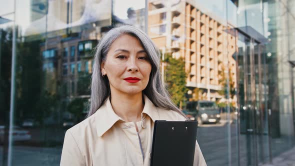 Asian Mature Lady is Holding Black Folder Looking at you While Standing Against Glass Facade of alt