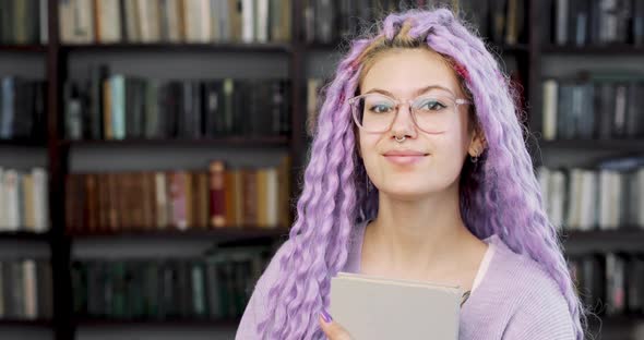 Portrait of a Young Woman in a Glasses with Long Colored Purple Hair Standing in the Library