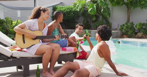Happy biracial woman playing guitar and singing with group of diverse friends at pool party alt