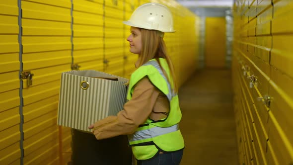 Serious Young Woman in Uniform and Hard Hat Walking Away with Box in Warehouse alt