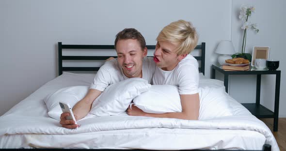 Homosexual Couple Hugging Looking at Smartphone Camera When Making Selfie Photos alt