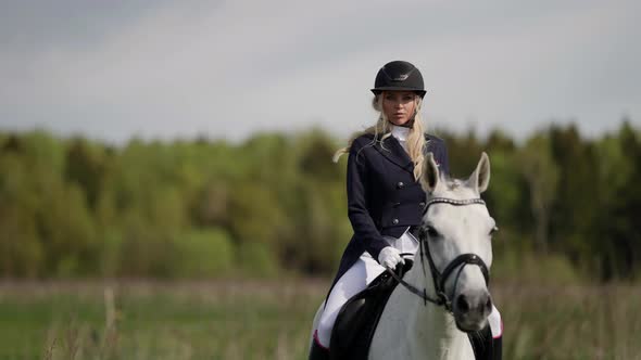 A Female Professional Jockey Riding a White Horse Gallops Across a Field alt