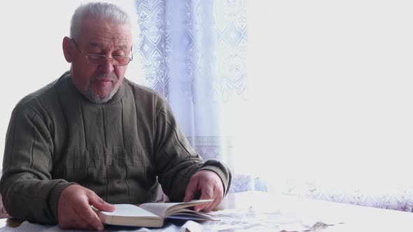 A Pensioner with Glasses Who Reads a Book While Sitting on a Chair in the House alt