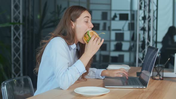 Young Businesswoman Eating Hamburger While Working on Laptop at the Office Table alt