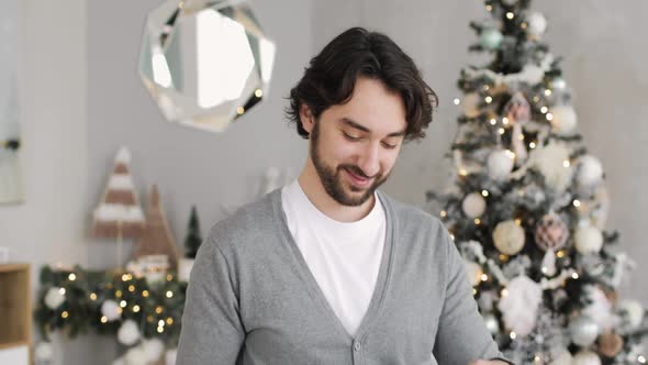 Young Happy Man Is Opening a Gift Box Near Christmas Tree at Home