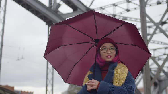 Cheerful Asian Woman Walking with Umbrella on Urban Bridge alt