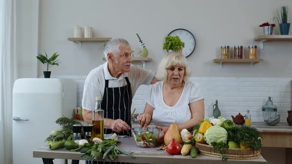 Elderly Grandparents in Kitchen. Funny Grandpa Joking on Grandma. Putting a Lettuce About Her Head alt