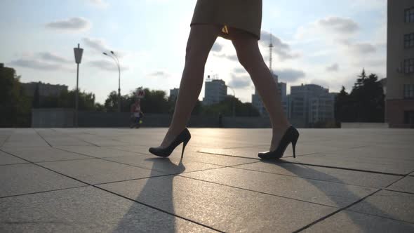 Slim Female Legs in Black Shoes on High Heels Walking on City Square at Sunset alt
