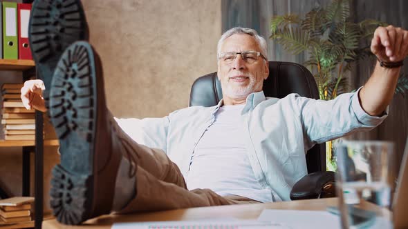 Grayhaired Man Putting Feet on Desk Next to Notebook and Smiling Leaning Back in Armchair Working at alt