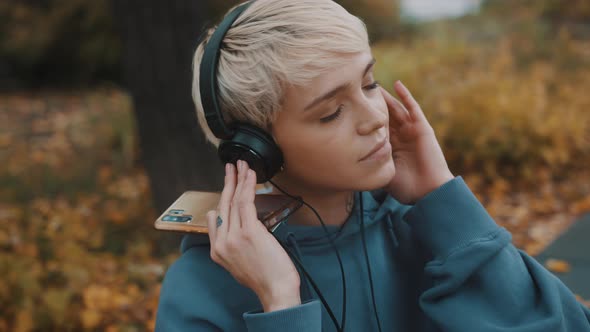 Young Blond Woman Listening To the Music From Her Smartphone in the Park Using the Headphones alt