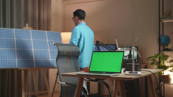 Asian Man Walks Into The Office Looking At Solar Cell That Is Next To The Green Screen Laptop alt