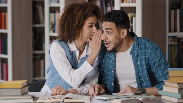 Two Students of Classmate Sitting at Desk in Lesson Doing Homework in Library Girl Whispers in Ear alt