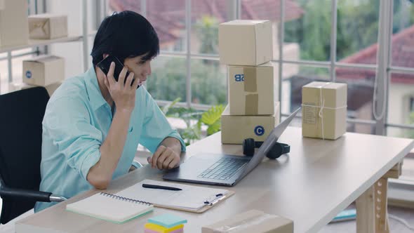 Asian man small business owner using laptop and checking parcel box and talking phone. alt
