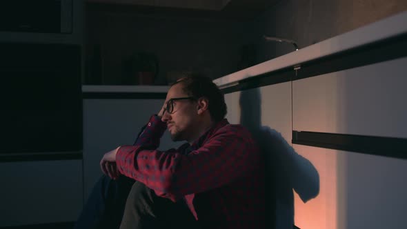 Young man with a thoughtful look sits on the kitchen floor at home and looks ahead alt