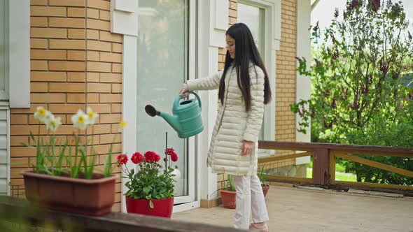 Wide Shot Side View of Young Slim Asian Woman Watering Flowers in Pot on Porch alt