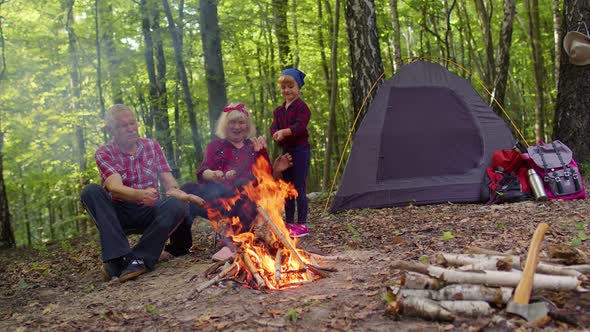 Senior Grandmother Grandfather with Granddaughter Cooking Frying Sausages Over Campfire in Wood alt