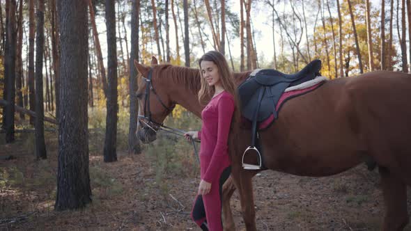 Side View of a Young Caucasian Girl in Pink Clothes Walking with Beautiful Brown Horse Between Trees alt