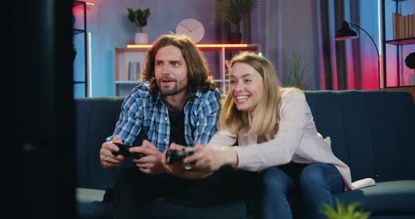 Couple Sitting on the Couch at Home in the Evening and Playing Videogames on TV Using Joysticks alt