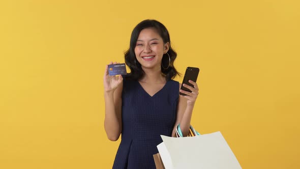Pretty smiling young Asian woman holding mobile phone and shopping bags showing credit card