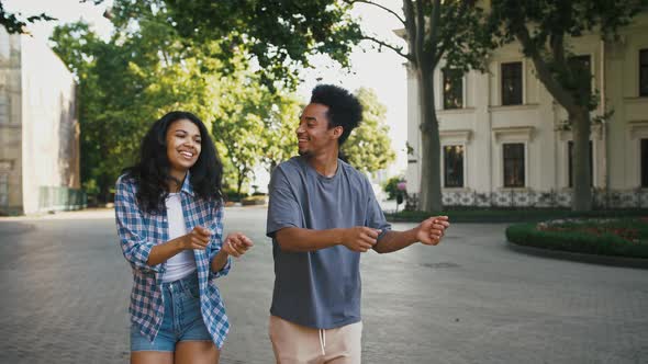 Ethnic Black Girl and Guy Smiling Dancing Synchronously While Walking By City Square alt