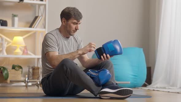 Serious Middle Aged Man Putting on Boxing Gloves Sitting on Exercise Mat alt
