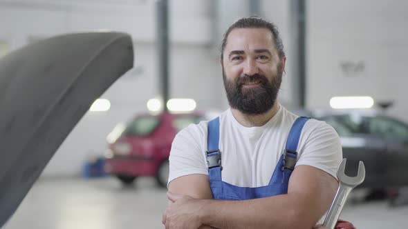 Portrait of a Smiling Bearded Mechanic Posing with a Wrench Standing at an Auto Repair Shop alt