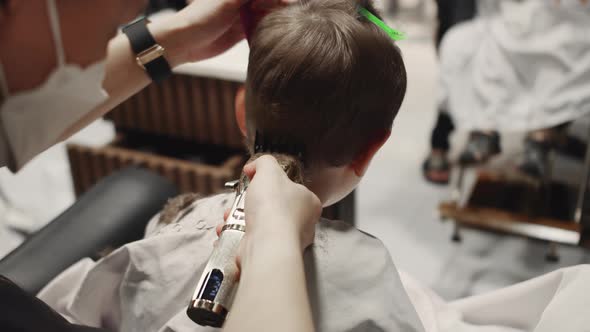 Fashionable Hairdresser Cuts a Child's Hair with a Clipper in a Barbershop alt