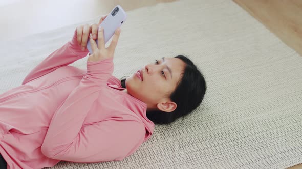 Young woman lying on floor using a smartphone after doing sport exercises indoor at home. alt