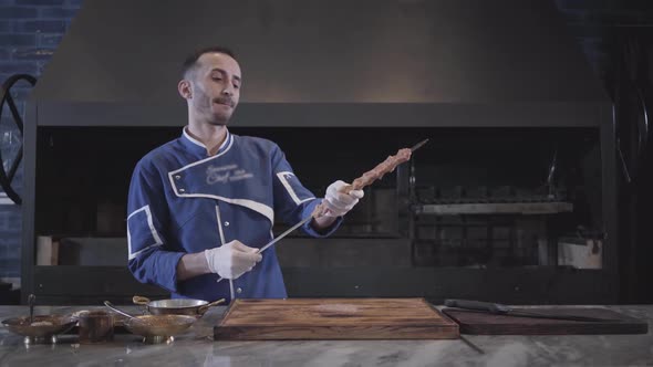 Skillful Man in Blue Chef Uniform Stringing Meat on a Skewer, Forming Lula Kebab in Modern alt