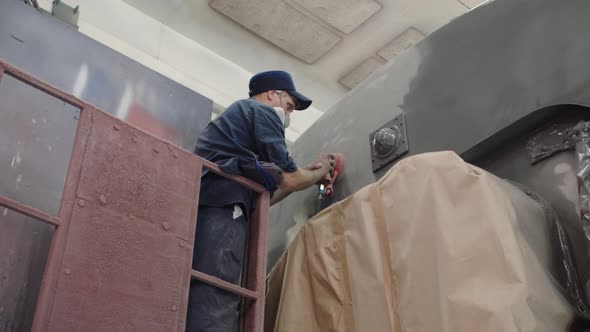 A Worker in a Cap and a Respirator Polishes the Paintwork of the Car Using a Pneumatic Grinder for