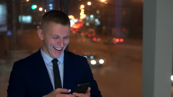 The Man-businessman Looks at the Smartphone Happy, Happy. Outside the Window, Against the Night City
