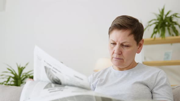 A Closeup View of a Beautiful Mature Woman is Reading a Newspaper Sitting on Sofa at Home in the alt