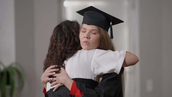 Young Female Graduate in Gown and Hat Hugging Woman with Dissatisfied Irritated Facial Expression alt